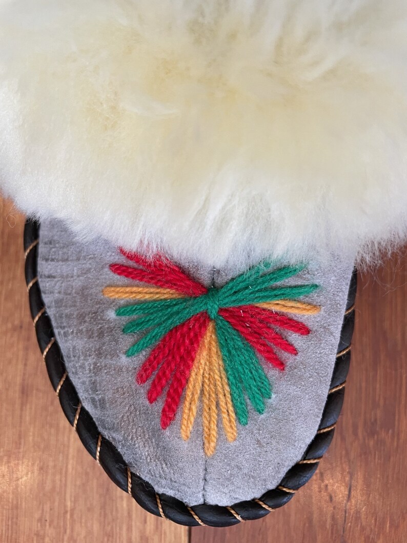 May include: Gray suede slipper with a black braided trim and a colorful embroidered design in red, yellow, and green.