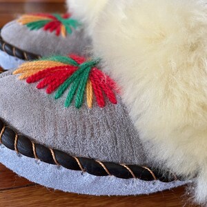May include: A pair of gray suede slippers with a black braided trim and colorful embroidered flowers. The slippers have a fluffy white lining.