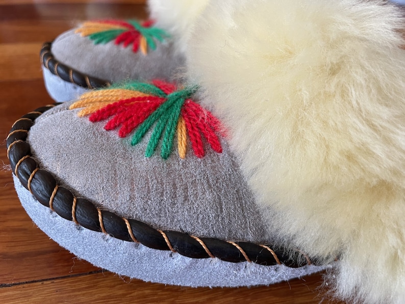 May include: A pair of gray suede slippers with a black braided trim and colorful embroidered flowers. The slippers have a fluffy white lining.