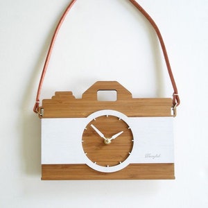 Puede incluir: Un reloj de madera con forma de cámara con una esfera blanca y agujas marrones. El reloj tiene una correa de cuero marrón y las palabras "Photolab" están impresas en la parte inferior.