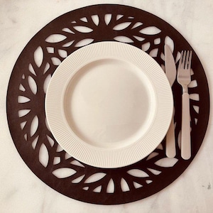 May include: A round, dark brown placemat with a leaf-like cutout design. A white plate with a ribbed edge sits in the center, accompanied by a knife and fork. This elegant table setting is perfect for a dining room.