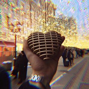May include: A wooden heart-shaped ornament with a grid pattern, held in a hand against a backdrop of twinkling lights.