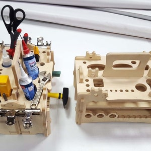 May include: Two wooden tool organizers with various compartments and slots for holding tools and supplies. The organizer on the left has a handle and is filled with tools, including scissors, pliers, glue bottles, and a wrench. The organizer on the right is empty and has a handle.