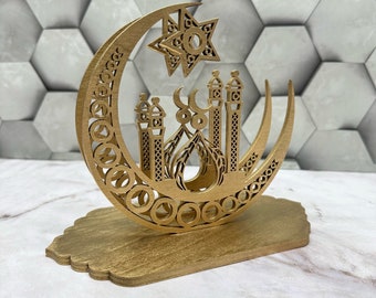 May include: A wooden decorative piece featuring a crescent moon, star, and mosque design. The intricate laser-cut details include geometric patterns and architectural elements. The piece is mounted on a rectangular base, all in a warm, natural wood tone.
