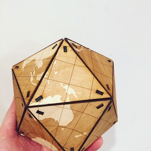 May include: A wooden geometric globe with a world map design. The globe is made of light brown wood and features a grid pattern. The continents are visible in a lighter shade. The globe is held in a hand.