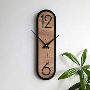 Può includere: Un orologio da parete ovale allungato con un quadrante in legno color marrone chiaro venato, racchiuso da una sottile cornice nera. L'orologio presenta grandi numeri neri per il 12 e il 6, con sottili lancette nere per ore, minuti e secondi.