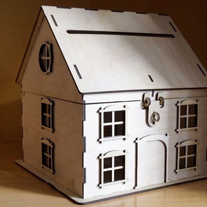May include: A wooden house-shaped box with a slot in the roof for cards or envelopes. The box has a hinged door and windows with black frames. The box is made of light wood and has a natural finish.