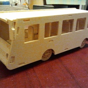 May include: A wooden model bus with a light brown finish. The bus has windows on the sides and a door on the front. The bus is sitting on a red surface.
