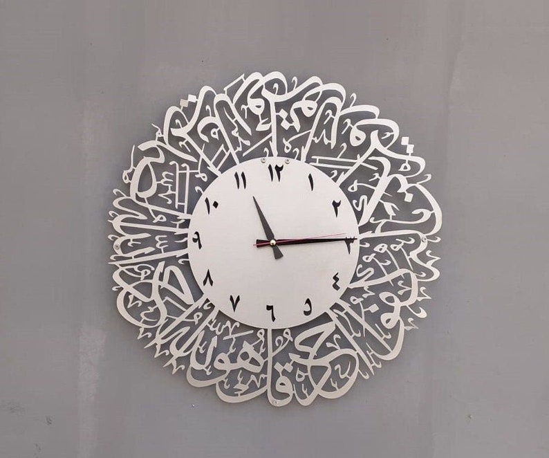May include: A white clock with Arabic calligraphy around the face. The clock has black hands and black Roman numeral markings.