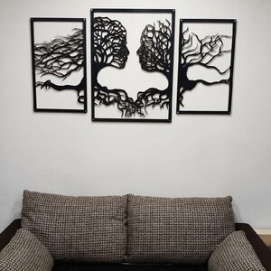 May include: Three black metal wall art pieces depicting two intertwined trees forming faces, with the middle piece showing the faces looking at each other. The art is displayed on a white wall above a brown couch.