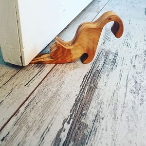 May include: A wooden doorstop in the form of a cat. The doorstop is made of light brown wood and is placed on a distressed, light-coloured wooden floor. It is positioned against a white door. The doorstop is approximately 15 cm long.