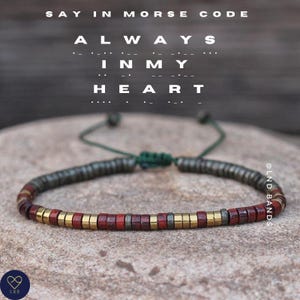 May include: A beaded bracelet with red, gold, and gray beads. The bracelet is adjustable with a green cord. The text "ALWAYS IN MY HEART" is written in Morse code above the bracelet.