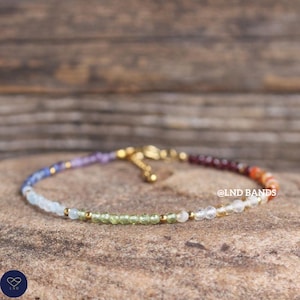May include: A multi-coloured beaded bracelet with a gold clasp. The bracelet features small, round gemstones in shades of blue, purple, green, orange, and red. The bracelet is displayed on a textured, brown surface.