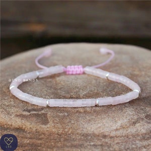 Könnte beinhalten: Ein rosa und silbernes Armband mit einem verschiebbaren Knotenschluss. Das Armband besteht aus kleinen, quadratischen rosa Steinen und silbernen Perlen. Das Armband liegt auf einer hellbraunen, strukturierten Oberfläche. @LND BANDS ist auf der Oberfläche geschrieben.