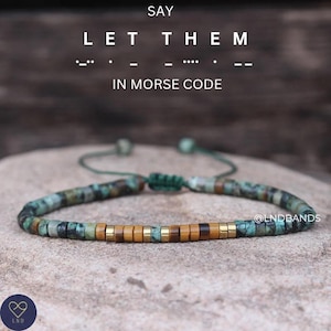 May include: A green, brown and gold beaded bracelet with a sliding knot closure. The bracelet has the words "LET THEM" written in Morse code.