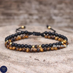 Puede incluir: Dos pulseras de cuentas. Una pulsera presenta cuentas negras mate y detalles dorados, mientras que la otra tiene cuentas de ojo de tigre y detalles dorados. Las pulseras están conectadas por un cordón negro. El texto "@LND BANDS" es visible.