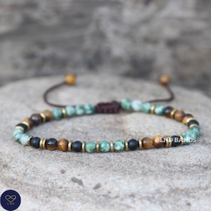 May include: A bracelet with a mix of green, brown, black, and gold beads. The bracelet has a sliding knot closure and is displayed on a stone surface. The text "@LND BANDS" is visible on the bracelet.
