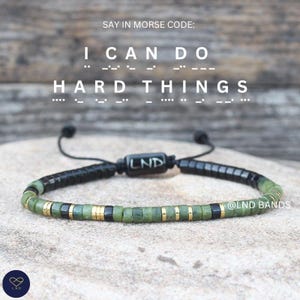 Puede incluir: Una pulsera de cuentas verdes, negras y doradas. La pulsera tiene un cordón negro ajustable y una cuenta negra central con las letras "LND". El texto "I CAN DO HARD THINGS" está encima de la pulsera.