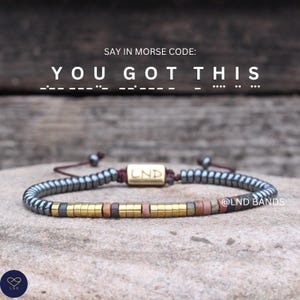Könnte beinhalten: Ein Armband mit einer Morsecode-Nachricht, die "YOU GOT THIS" sagt. Das Armband hat eine Mischung aus metallischen Perlen in Grau-, Gold- und Brauntönen. Der Markenname "LND" ist auf einem goldenen Balken zu sehen.