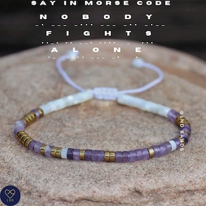 Könnte beinhalten: Ein violettes und weißes Perlenarmband mit goldenen Akzenten. Das Armband ist mit einer weißen Schnur gebunden und trägt den Text "NOBODY FIGHTS ALONE" in Morsecode.