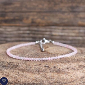 Op de afbeelding: Een delicate roze kralenarmband met een zilveren sluiting. De armband is gemaakt van kleine, ronde, lichtroze kralen. De achtergrond is een natuurlijk, getextureerd oppervlak. De tekst "@LND.BANDS" is zichtbaar.