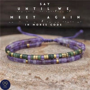 May include: A double-layered bracelet with purple and green beads and gold accents. The bracelet is tied with a purple cord and says "SAY UNTIL WE MEET AGAIN" in Morse code.