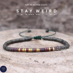 Op de afbeelding: Een kralenarmband met de tekst "STAY WEIRD" erboven. De armband heeft donkergrijze, rode en gouden kralen. De armband ligt op een neutraal getinte ondergrond. De merknaam "@LND BANDS" is zichtbaar.