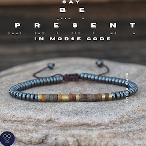 May include: A black beaded bracelet with a colorful stone accent. The bracelet is tied with a brown cord and has a sliding knot for adjusting the fit. The bracelet is on a gray textured surface. The text "SAY BE PRESENT IN MORSE CODE" is above the bracelet.