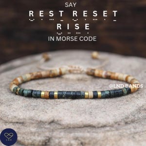 May include: Beaded bracelet with a mix of earth-toned beads, including green, brown, and gold accents. The bracelet is set against a textured stone surface. Text reads "SAY REST RESET RISE IN MORSE CODE".