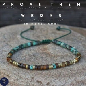 Pode incluir: Um bracelete de contas com contas turquesa, castanhas e douradas. O bracelete tem um cordão verde e um fecho de nó deslizante. O texto "PROVE THEM WRONG IN MORSE CODE" é exibido acima do bracelete.