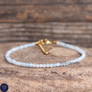Op de afbeelding: Fijn kralenarmbandje met kleine, lichtblauwe en witte edelstenen. De armband heeft een goudkleurige sluiting en wordt getoond op een getextureerd, bruin oppervlak. De tekst "@LND BANDS" is zichtbaar.