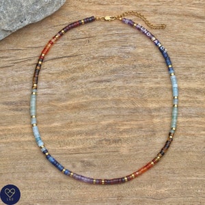 May include: A multi-coloured beaded necklace with a gold clasp. The beads are arranged in a gradient pattern, starting with red and orange at the bottom and transitioning to blue and purple at the top. The necklace is on a wooden surface.