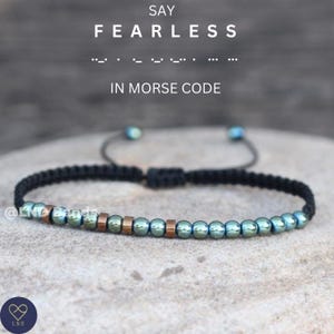 Może przedstawiać: Bransoletka z czarnego sznurka z zielonymi i miedzianymi koralikami. Bransoletka zakodowana jest w alfabecie Morse'a jako "FEARLESS".