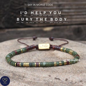 May include: A beaded bracelet with green, maroon, and gold accents. The bracelet features the text "I'D HELP YOU BURY THE BODY" in Morse code. The bracelet has a gold clasp with the letters "LND" on it. The bracelet is on a stone surface.