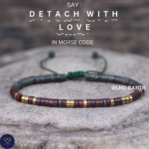 Puede incluir: Una pulsera de cuentas con el mensaje "DETACH WITH LOVE" en código Morse. La pulsera presenta cuentas de color rojo oscuro y dorado, con un cordón verde ajustable. La imagen está sobre un fondo de piedra.
