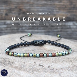 Op de afbeelding: Zwart kralenarmband met een mix van koperen en iriserende kralen. De armband heeft een schuifknoopsluiting en een kleine zwarte rechthoekige kraal met "LND". Bovenstaande tekst luidt "UNBREAKABLE" in morsecode.