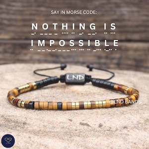 Puede incluir: Una pulsera con cuentas cilíndricas marrones, negras y doradas y una cuenta rectangular central negra marcada con LND. El texto SAY IN MORSE CODE: NOTHING IS IMPOSSIBLE aparece arriba con los puntos y rayas del código Morse.