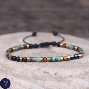 May include: A bracelet with a mix of turquoise, brown, black, and gold-coloured beads. The bracelet is adjustable with a black cord. The bracelet is displayed on a neutral-toned surface. The bracelet has the text "@ND Bands".