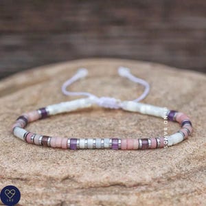 May include: A delicate bracelet featuring pink, purple, and white beads. The bracelet is made with a white cord and has a sliding knot for adjusting the size.