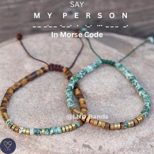 Peut inclure: Deux bracelets en perles avec des perles brunes et vertes. Les bracelets sont liés ensemble par un cordon brun. Le texte "SAY MY PERSON In Morse Code" est au-dessus des bracelets.