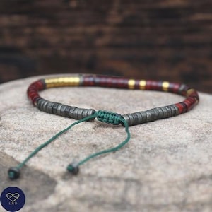 May include: A handmade bracelet featuring a combination of dark gray, red, and gold-colored beads. The bracelet is strung on a green cord with a sliding knot closure. The beads are cylindrical and arranged in a pattern.