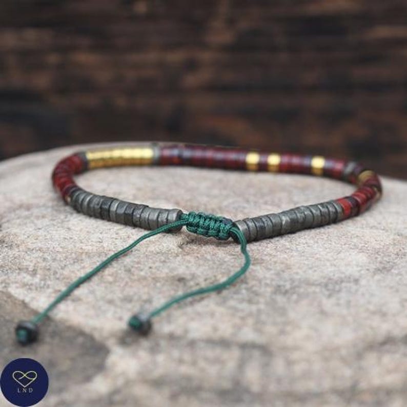 May include: A handmade bracelet featuring a combination of dark gray, red, and gold-colored beads. The bracelet is strung on a green cord with a sliding knot closure. The beads are cylindrical and arranged in a pattern.