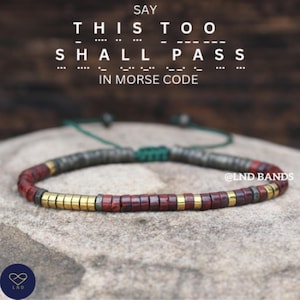 Op de afbeelding: Een armband met de tekst "THIS TOO SHALL PASS" in morsecode. De armband heeft rode, gouden en grijze kralen en een groen koord. De tekst "SAY THIS TOO SHALL PASS IN MORSE CODE" staat bovenaan.