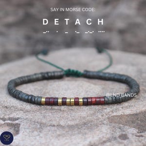 May include: A beaded bracelet with the word "DETACH" in Morse code. The bracelet features dark gray beads, with accents of gold and reddish-brown beads. The bracelet is on a stone surface, with a green cord.
