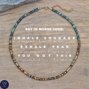 Może przedstawiać: Naszyjnik z turkusowych i złotych koralików z zapięciem. Naszyjnik zawiera napis "INHALE COURAGE, EXHALE FEAR, YOU GOT THIS" w alfabecie Morse'a.