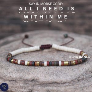 May include: A multi-coloured beaded bracelet with a cream-coloured cord. The beads are red, brown, gold, and white. The text "ALL I NEED IS WITHIN ME" is above the bracelet in Morse code.