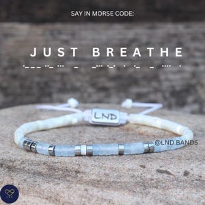 May include: A light blue and white beaded bracelet with silver accents. The bracelet has the words "JUST BREATHE" above it, written in Morse code. The bracelet is on a stone surface. The brand name "LND" is visible on the bracelet and in the bottom left corner.