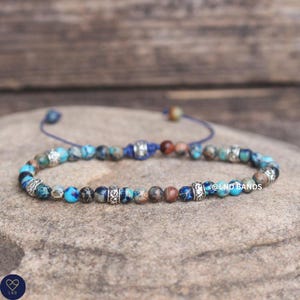 May include: A beaded bracelet featuring round, multi-coloured stones in shades of blue, brown, and grey, interspersed with ornate silver-toned metal spacers. The bracelet includes a blue cord closure and displays the text @LND BANDS on the side.