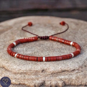 May include: Red and silver beaded bracelet with a brown cord and adjustable closure. The bracelet has the text "@LND BANDS" on it.