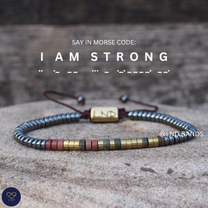 May include: A bracelet featuring metallic grey, red, and gold-coloured cylindrical beads on a dark cord. A central gold-toned rectangular bead is engraved with the letters LND. Text above reads SAY IN MORSE CODE: I AM STRONG followed by Morse code dots and dashes.
